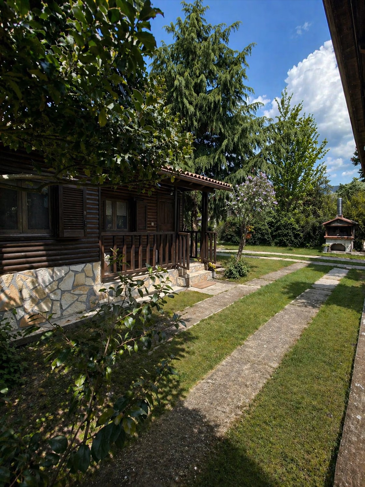 The Cozy Chalet's veranda running along the side of the house, with stone paths crossing the lawn towards a stone oven.