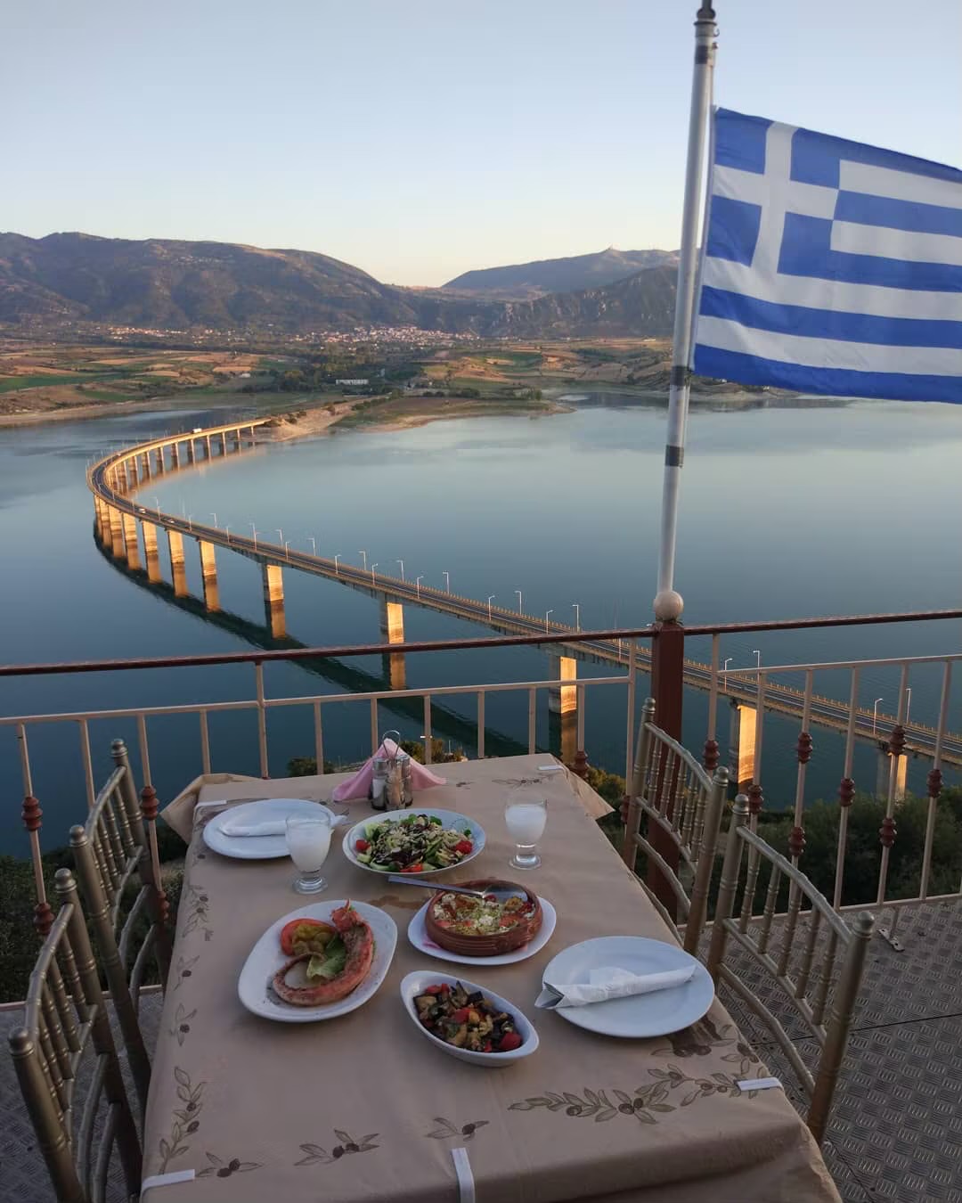 Ένα τραπέζι με μεζέδες σε βεράντα πάνω από τη λίμνη, με μια ελληνική σημαία δίπλα το σούρουπο.