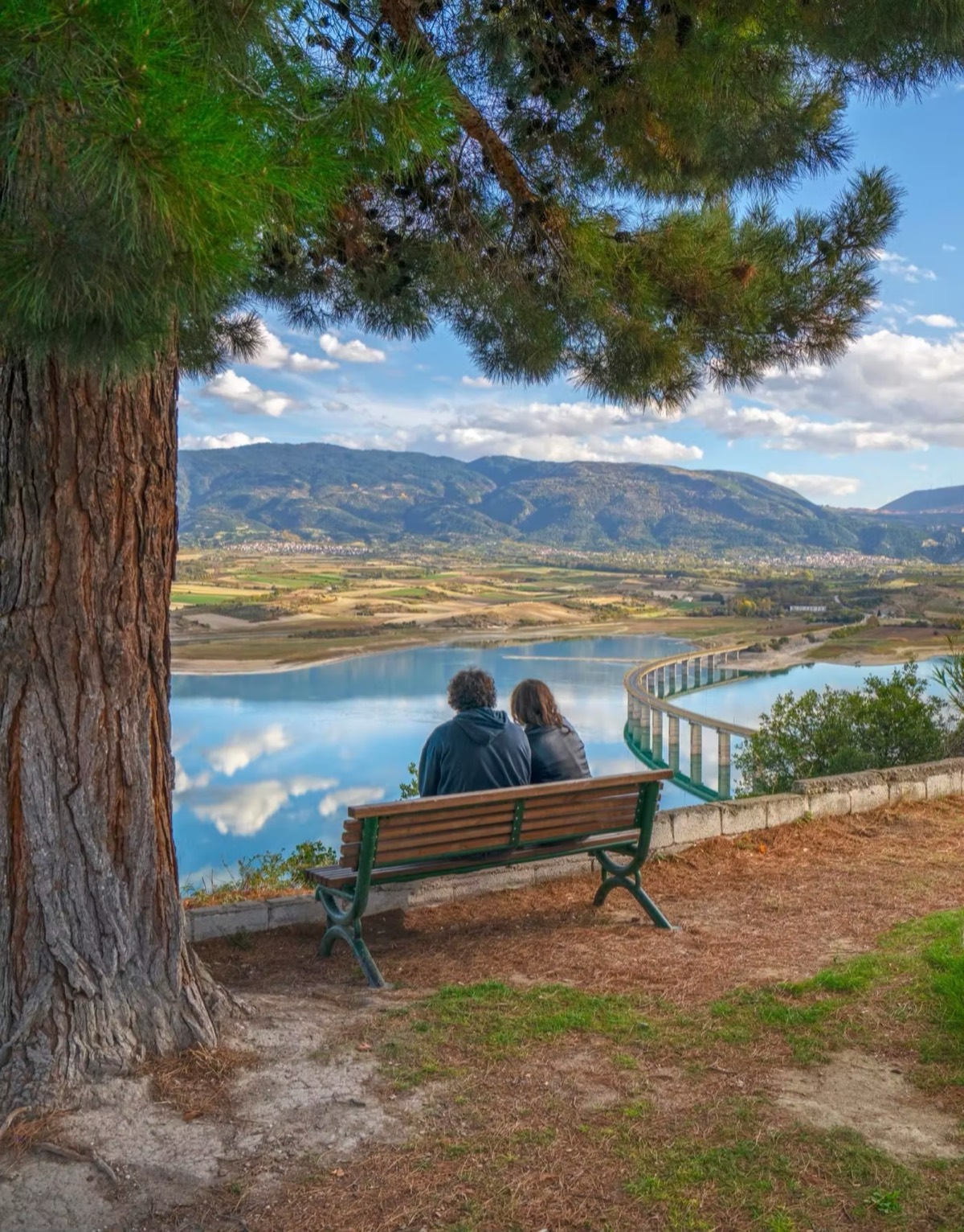 Δύο άνθρωποι σε ένα παγκάκι κάτω από ένα πεύκο, κοιτάζοντας τη λίμνη.