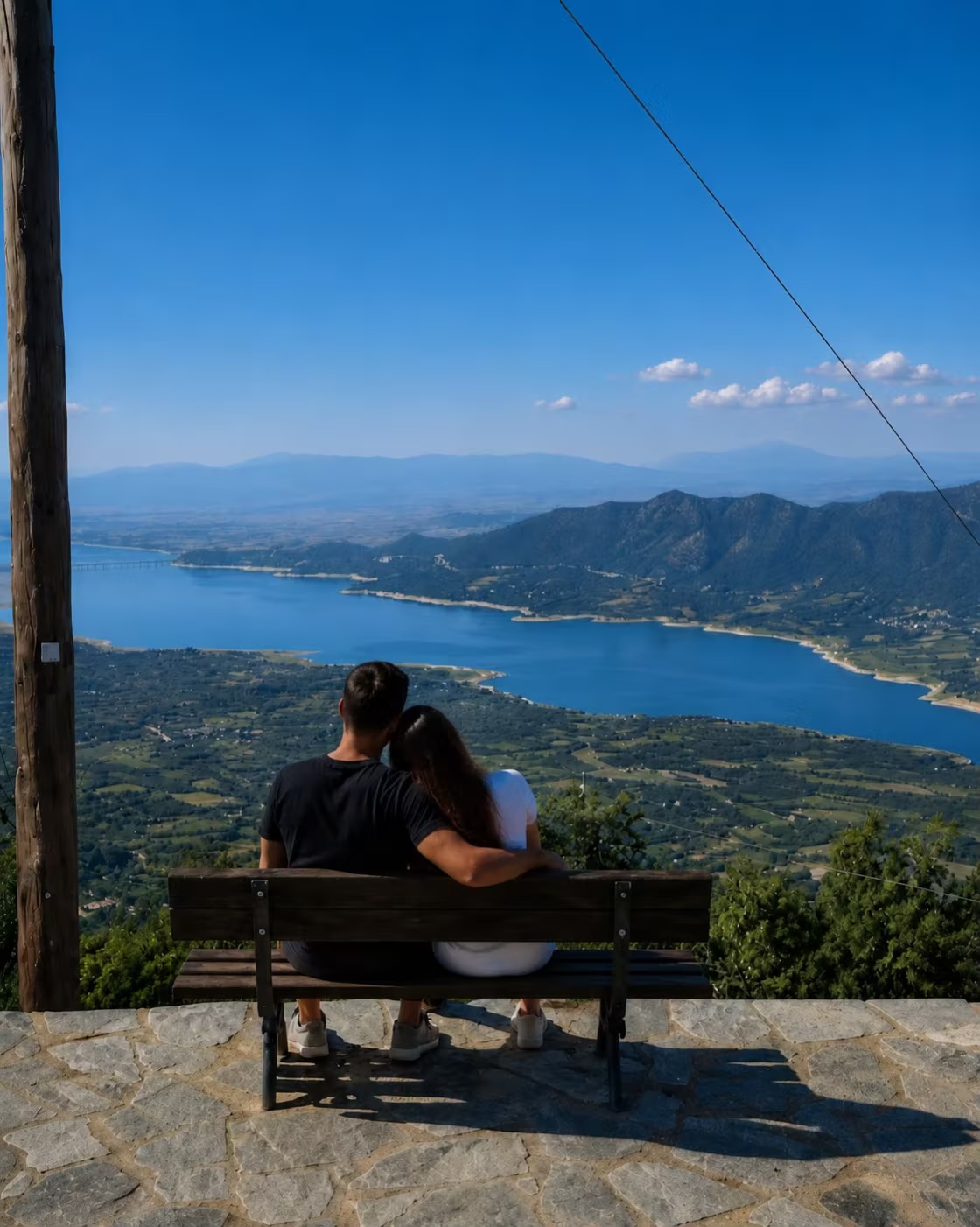 Ένα ζευγάρι σε παγκάκι σε ορεινό σημείο θέας, με τη λίμνη Πολυφύτου να απλώνεται πολύ πιο κάτω.