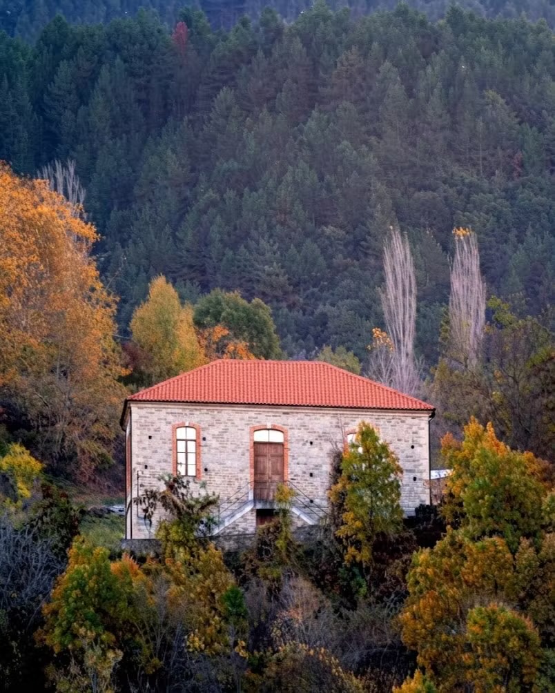 Ένα πέτρινο σπίτι με κεραμοσκεπή ανάμεσα σε φθινοπωρινά δέντρα, με πευκοδάσος να υψώνεται πίσω του.