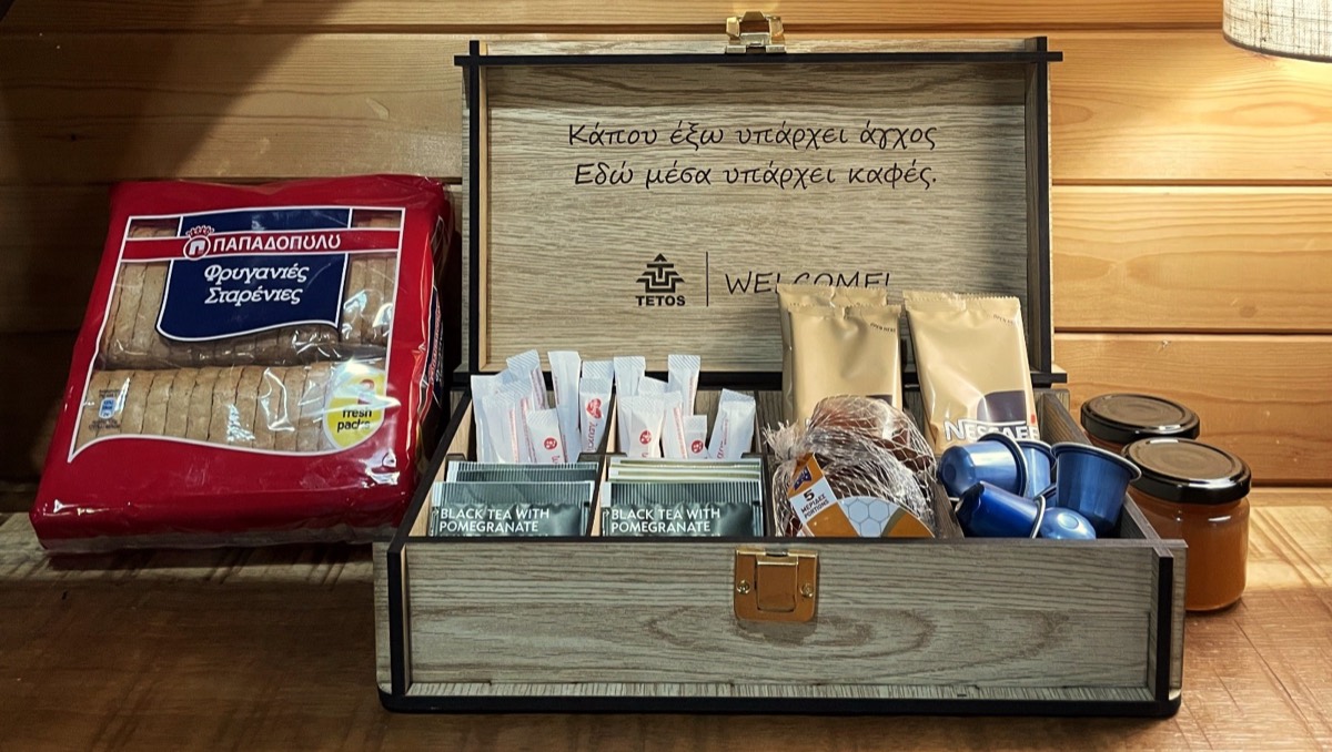 Ένα ξύλινο welcome box ανοιχτό πάνω σε τραπέζι, με χαραγμένο στο καπάκι &ldquo;Κάπου έξω υπάρχει άγχος. Εδώ μέσα υπάρχει καφές.&rdquo; πάνω από το σήμα της TETOS &mdash; μέσα, τσάι, ζάχαρη σε φακελάκια, στιγμιαίος καφές, κάψουλες Nespresso, μια σακούλα με μερίδες μέλι και δύο βαζάκια, και δίπλα ένα πακέτο φρυγανιές.